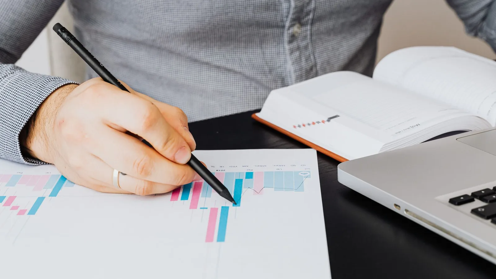 A person marking a printed bar chart with a pencil, beside an open notebook and a laptop
