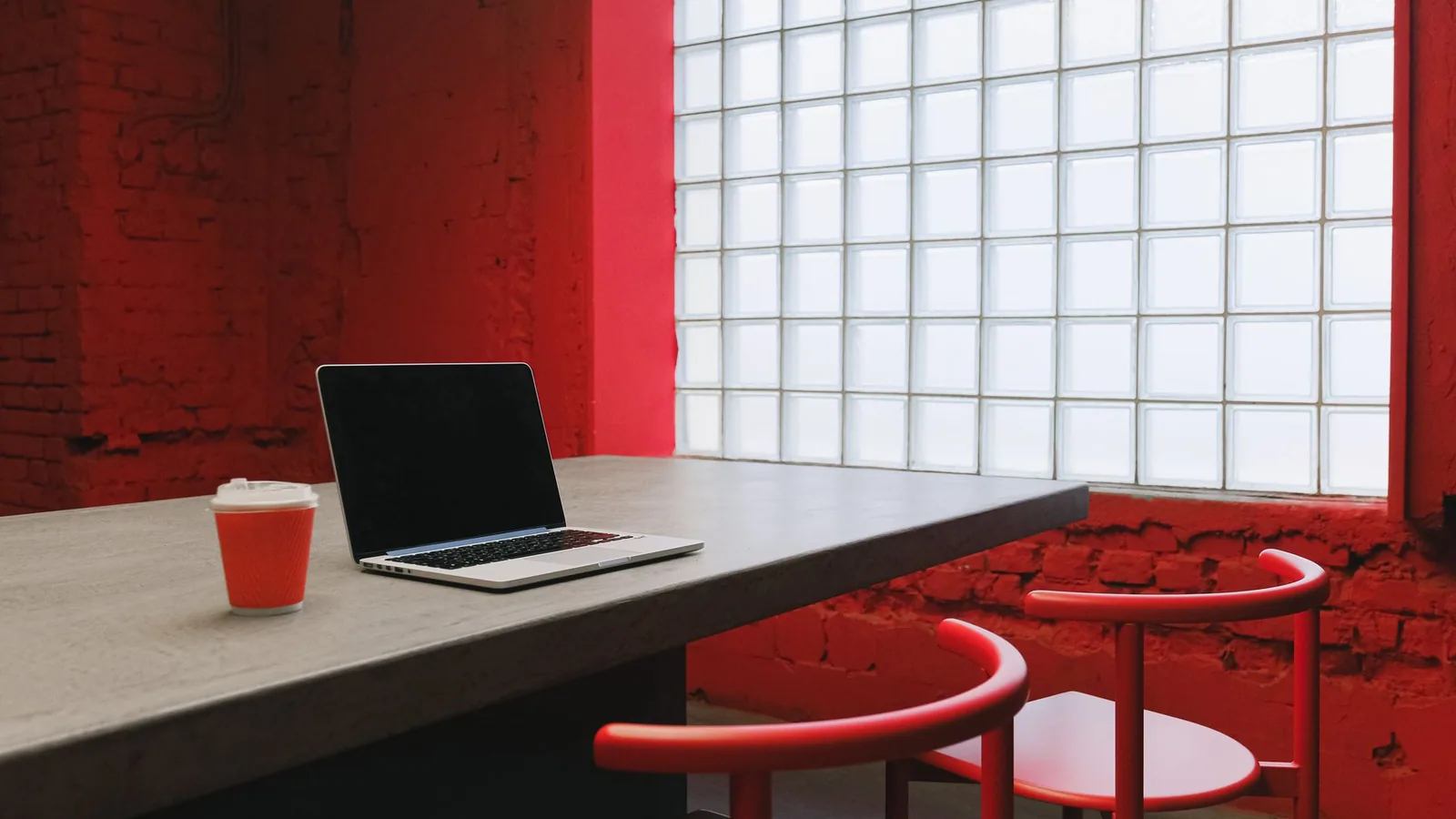 An open laptop and a takeaway coffee cup on a grey table in a red painted room, with red chairs and a glass block window