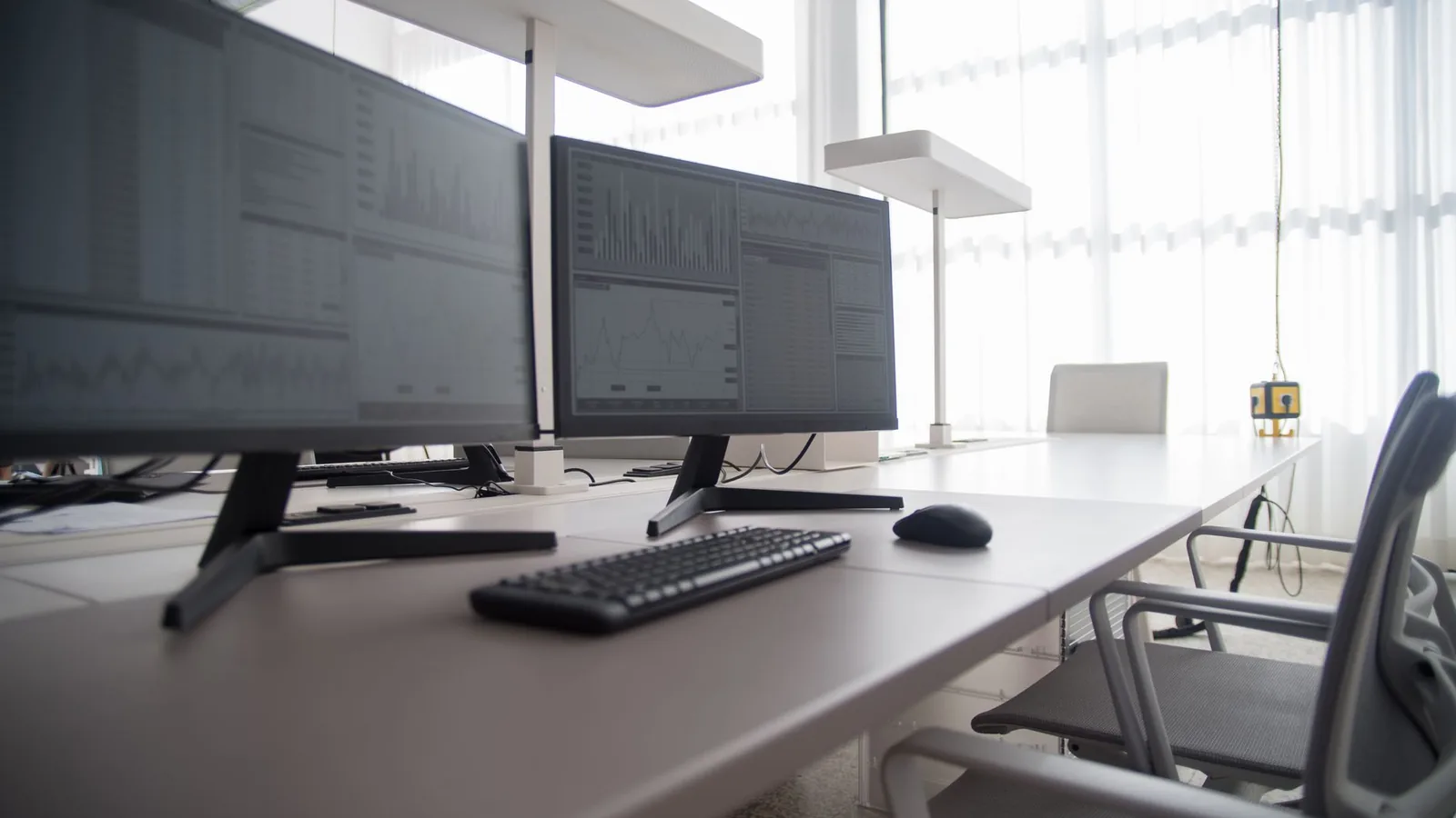 Two monitors showing spreadsheets and charts on a white desk with a keyboard and mouse in an empty office