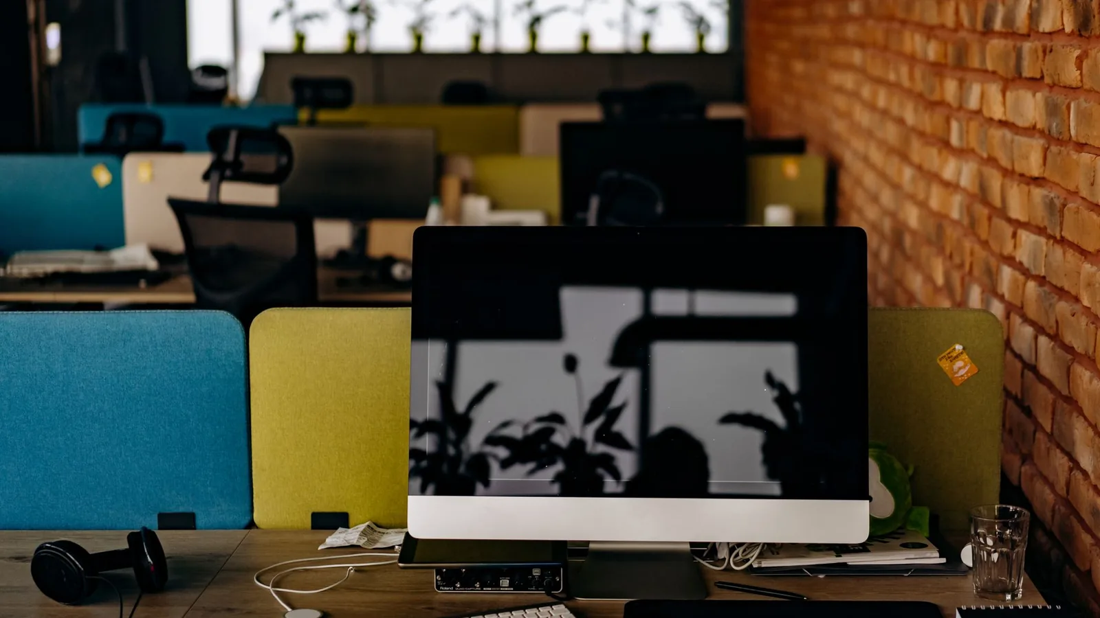A computer monitor on a desk in an empty open plan office, with coloured desk dividers and a brick wall