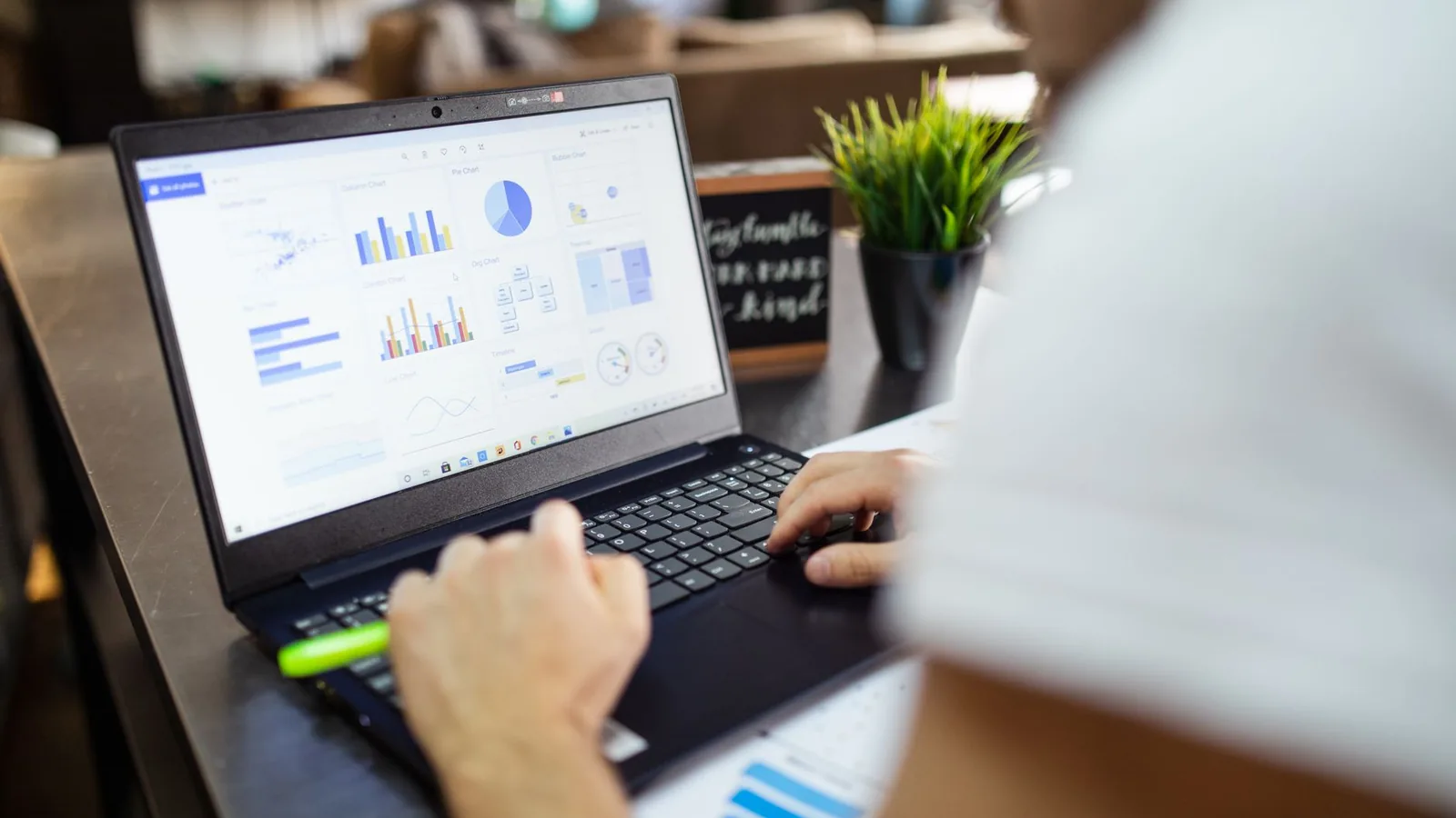 A person typing on a laptop showing bar charts and a pie chart, beside a small potted plant on a wooden desk
