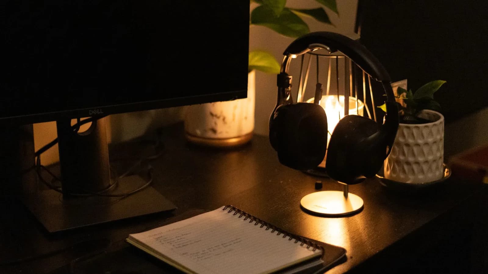 A desk at night lit by a small lamp, with a monitor, headphones on a stand and an open handwritten notebook